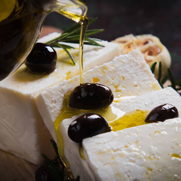 Nærbillede af en kande, der hælder olivenolie over frisk feta og oliven.